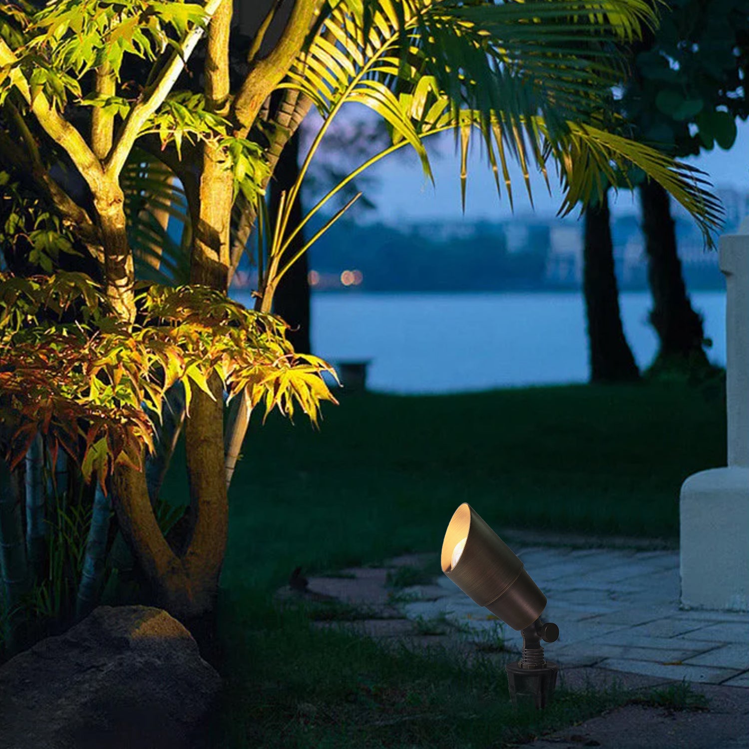 12V LED Low Voltage Solid Brass Outdoor Light illuminating a landscaped area with trees and foliage at dusk, highlighting greenery beside a stone pathway.