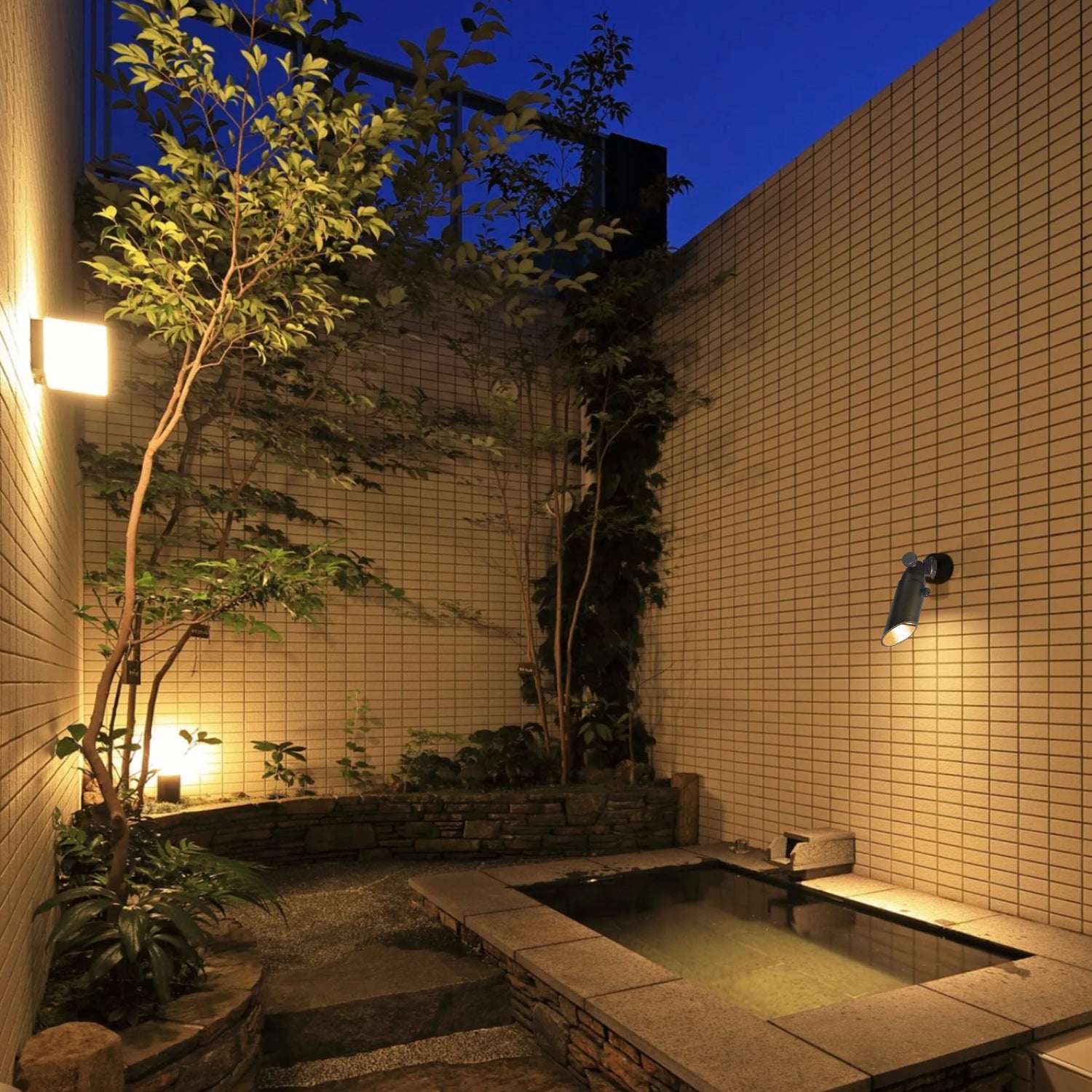 Outdoor garden at night illuminated by wall-mounted lights and a mini brass low voltage spotlight, highlighting tiled walls, plants, and a small garden area with trees.