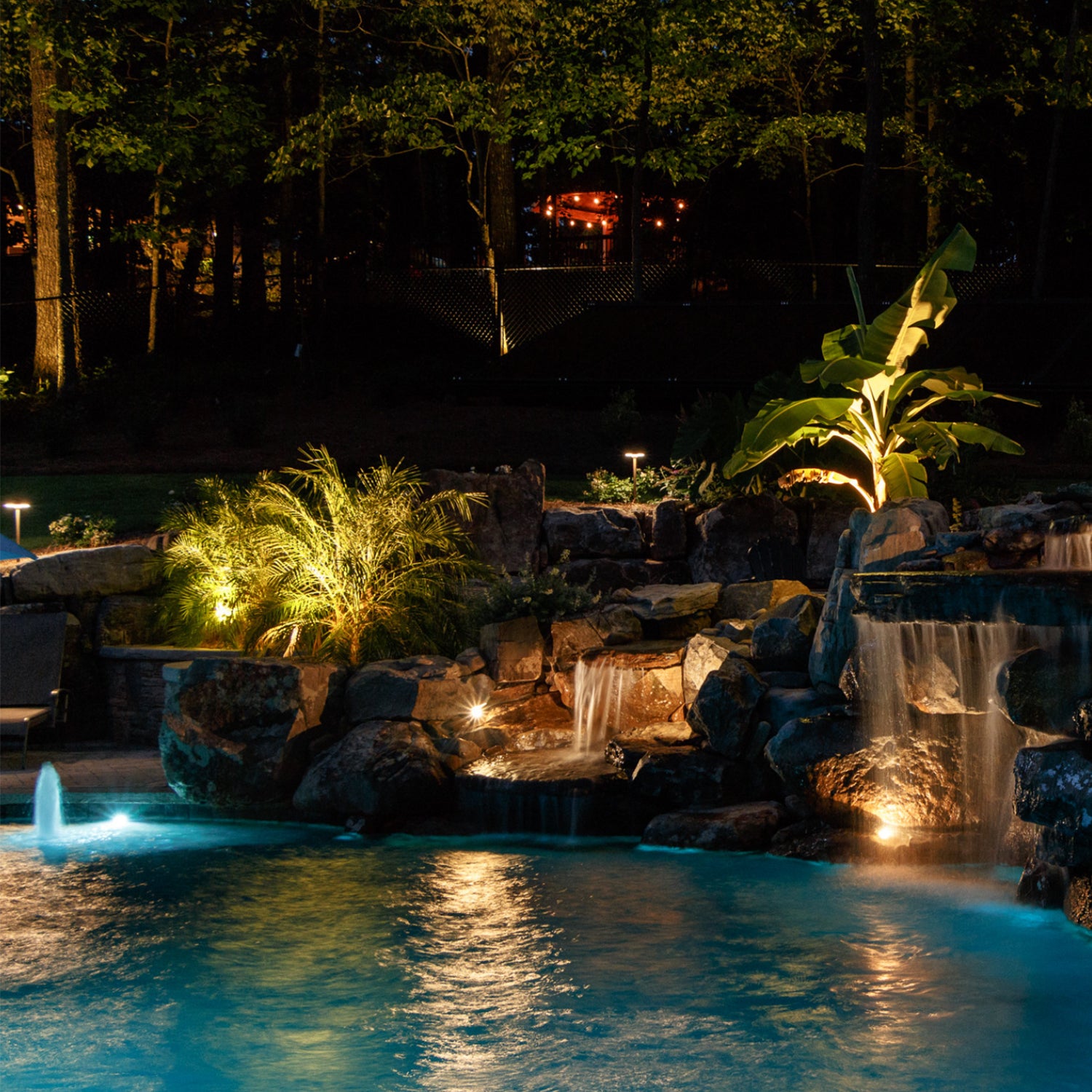 Night view of a backyard pool with waterfall feature illuminated by COLOER LED landscape lighting, showcasing underwater lights and garden spotlights enhancing the plants and rocks.