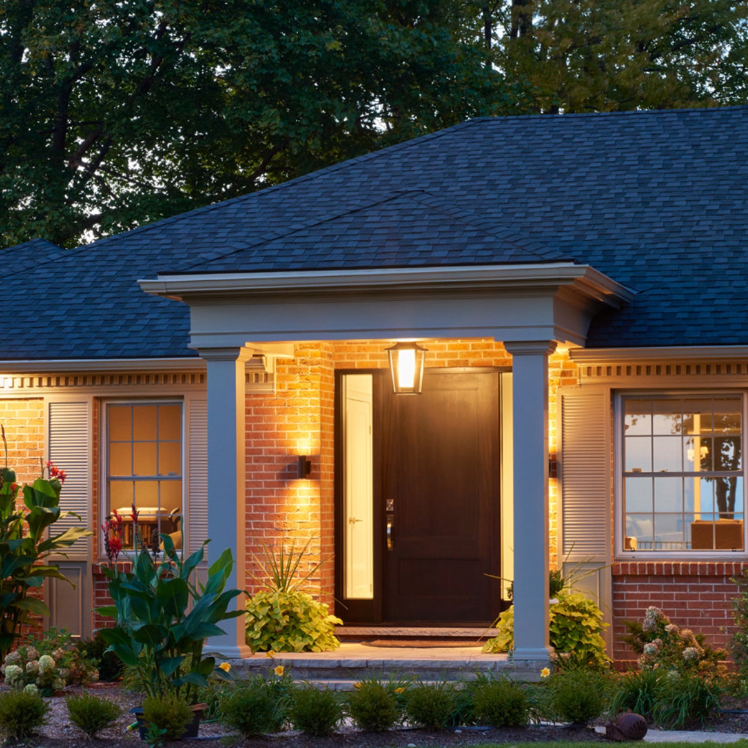 Brick house with a well-lit front entrance featuring a black door, columns, and brass wall sconces on either side of the door.