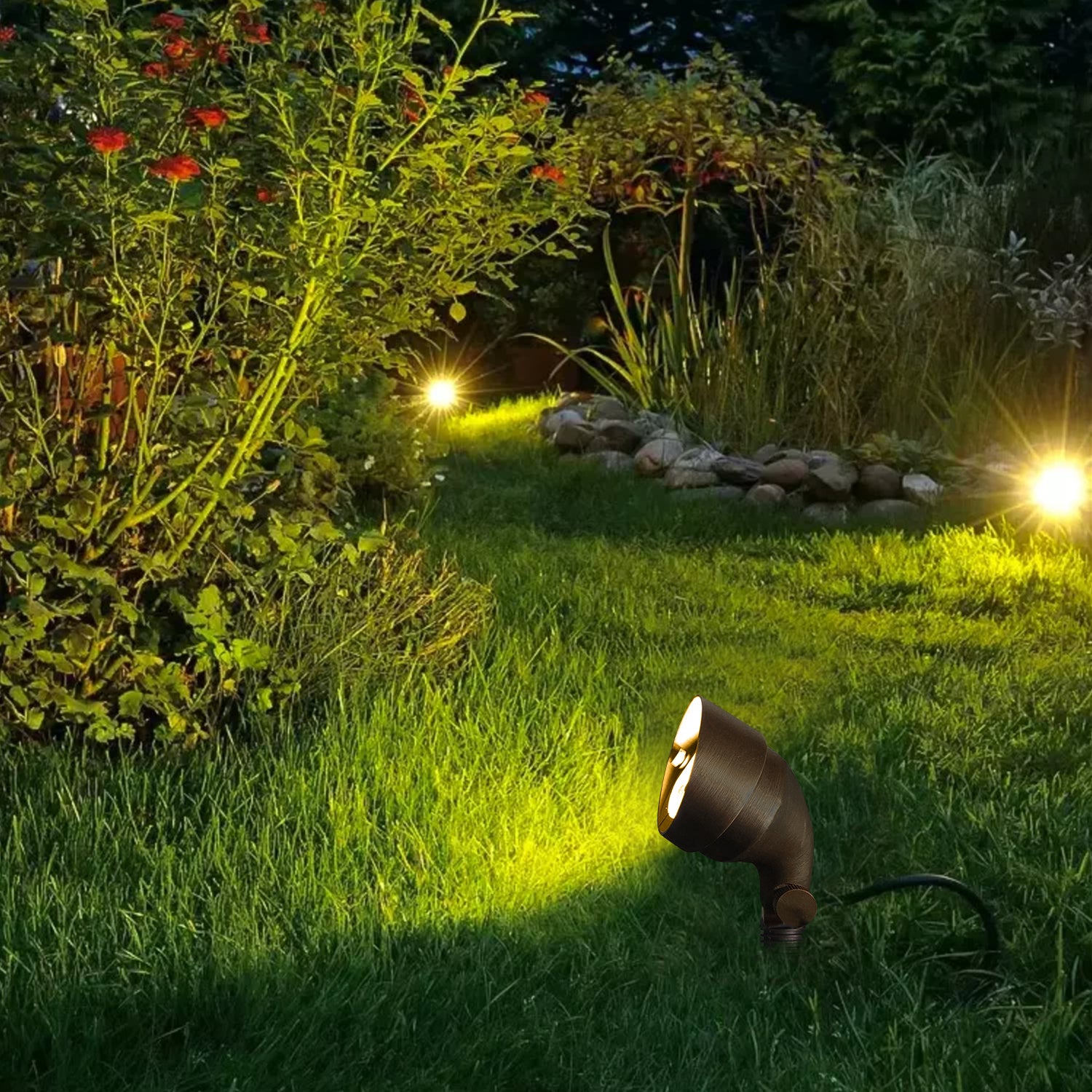 Garden illuminated at night with brass landscape flood lights highlighting plants and a rock feature, providing a warm, ambient glow