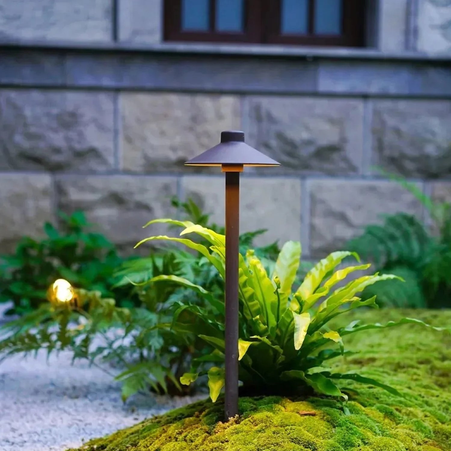 12V low voltage brass garden path light COP602B illuminating a garden with greenery and plants, with a stone wall and window in the background