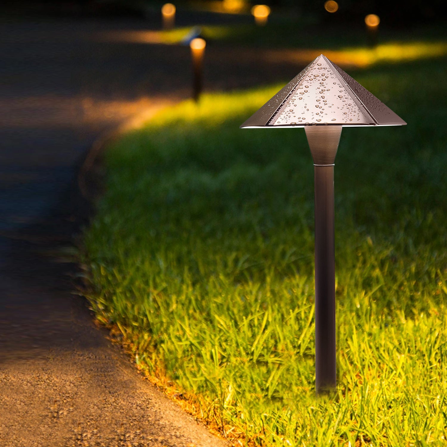 Brass low voltage pathway light illuminating a driveway and yard at night with other similar lights in the background